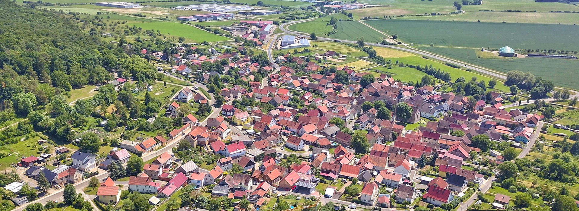 Weite Luftansicht von grünen Feldern, Industriegebäuden und einem Dorf vor einem bewaldeten Hintergrund.